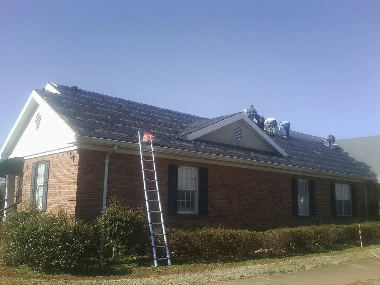 Crew tearing shingles off a brick ranch roof with a ladder set — Metal Roof Repair work by our crew in Vale